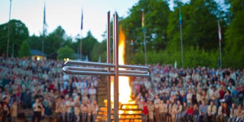 Im Hintergrund eines Kreuzes brennt ein Lagerfeuer mit vielen Menschen drumrum